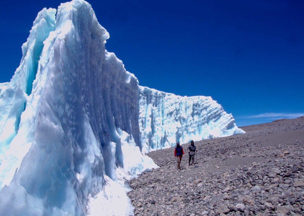 Two hikers walk past an ice wall on top of the Kilimanjaro.