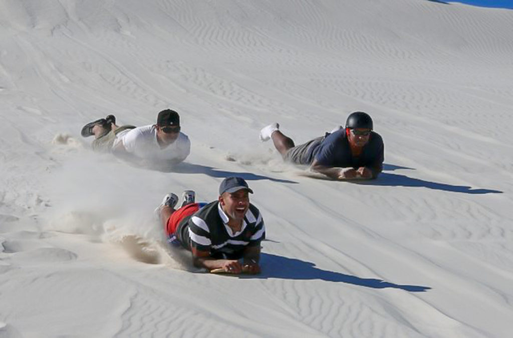 Drie mannen sandboarden liggend op hun buik van de witte duinen af.