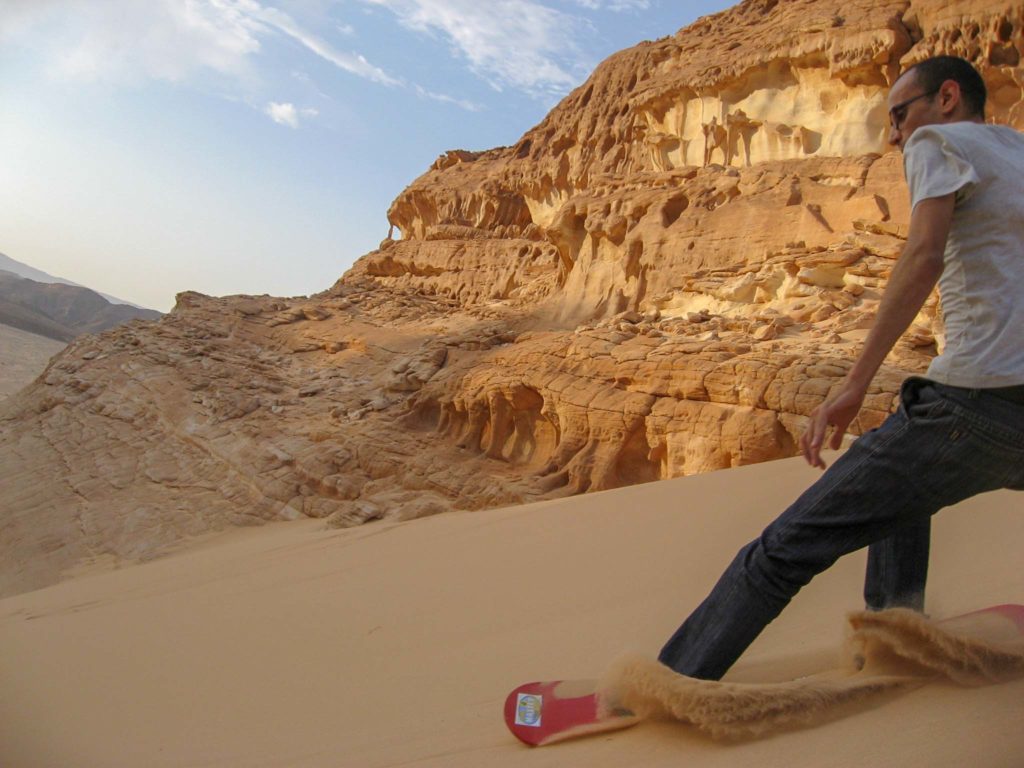 Een sandboarder remt af terwijl we op de achtergrond de oranje rotsen van de Sinaï woestijn zien.