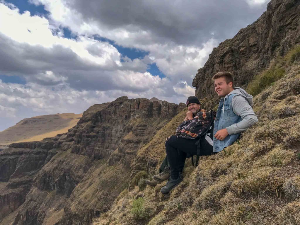 Twee mannen zitten op de steile rand van Lesotho. Ze kijken in de verte naar de Drakensbergen. 