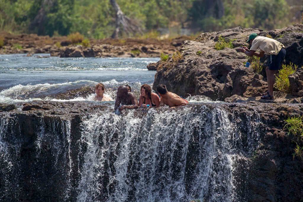 Gezien vanaf de Zimbabwaanse kant leunen drie mensen op de rand van Devils Pool terwijl een ander achter hen zwemt