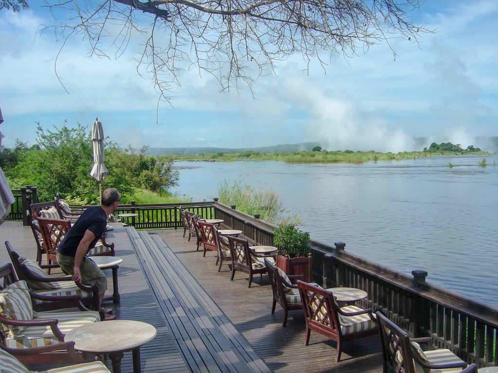 Een man gaat zitten op de veranda van the Royal Livingston Hotel terwijl hij kijkt naar het opstuivende water langs de rivier