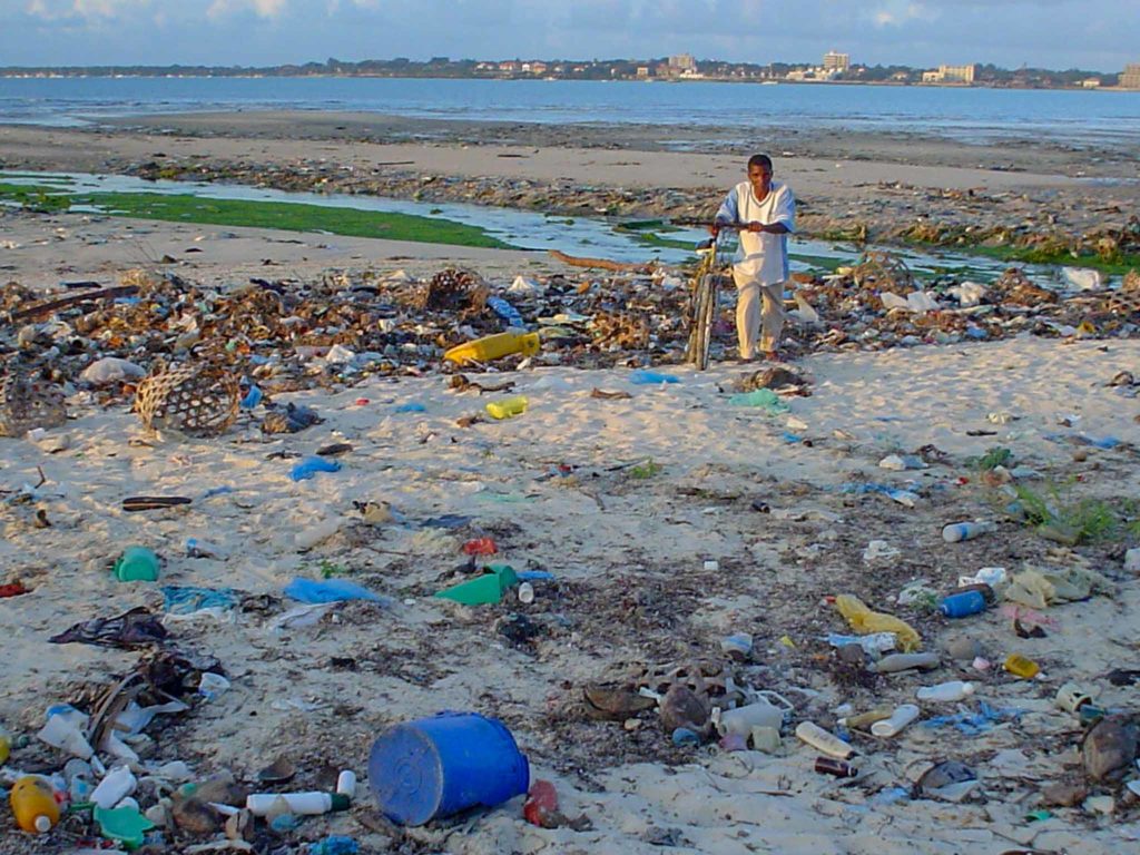 Een man loopt over het strand in Dar el Salaam met allerlei weggegooid plastic om hem heen.  