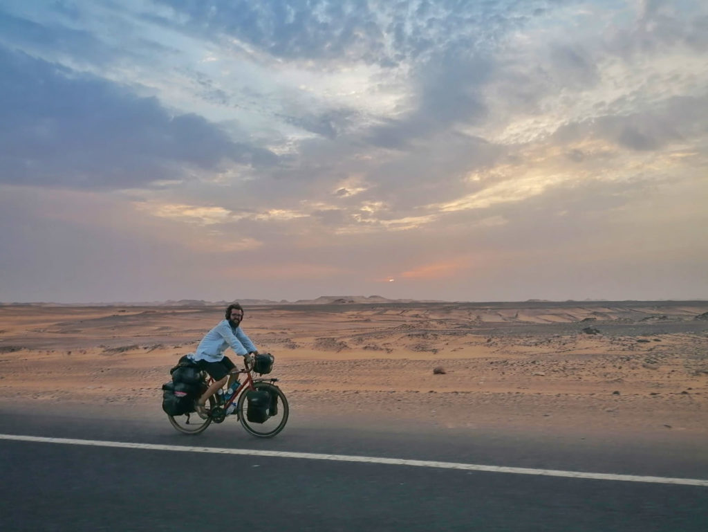 Koen fietst op een geasfalteerde weg met de donker oranje woestijn achter hem. In de verte begint de zon ook onder te gaan.