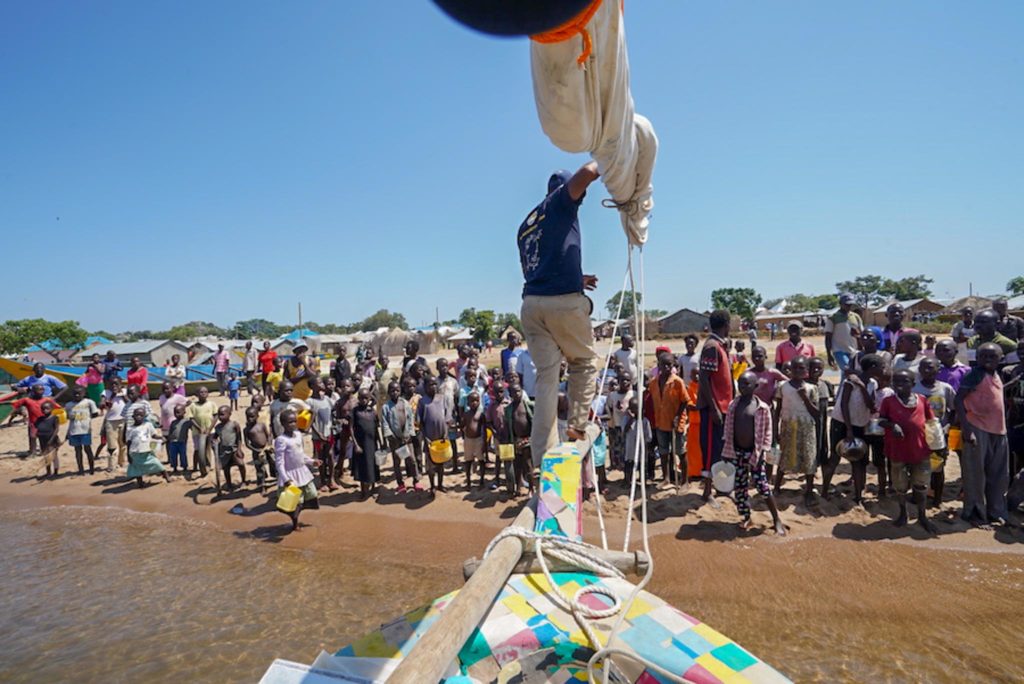 The nose of the Flipflopi reaches a shore with tens of people wainting for them.